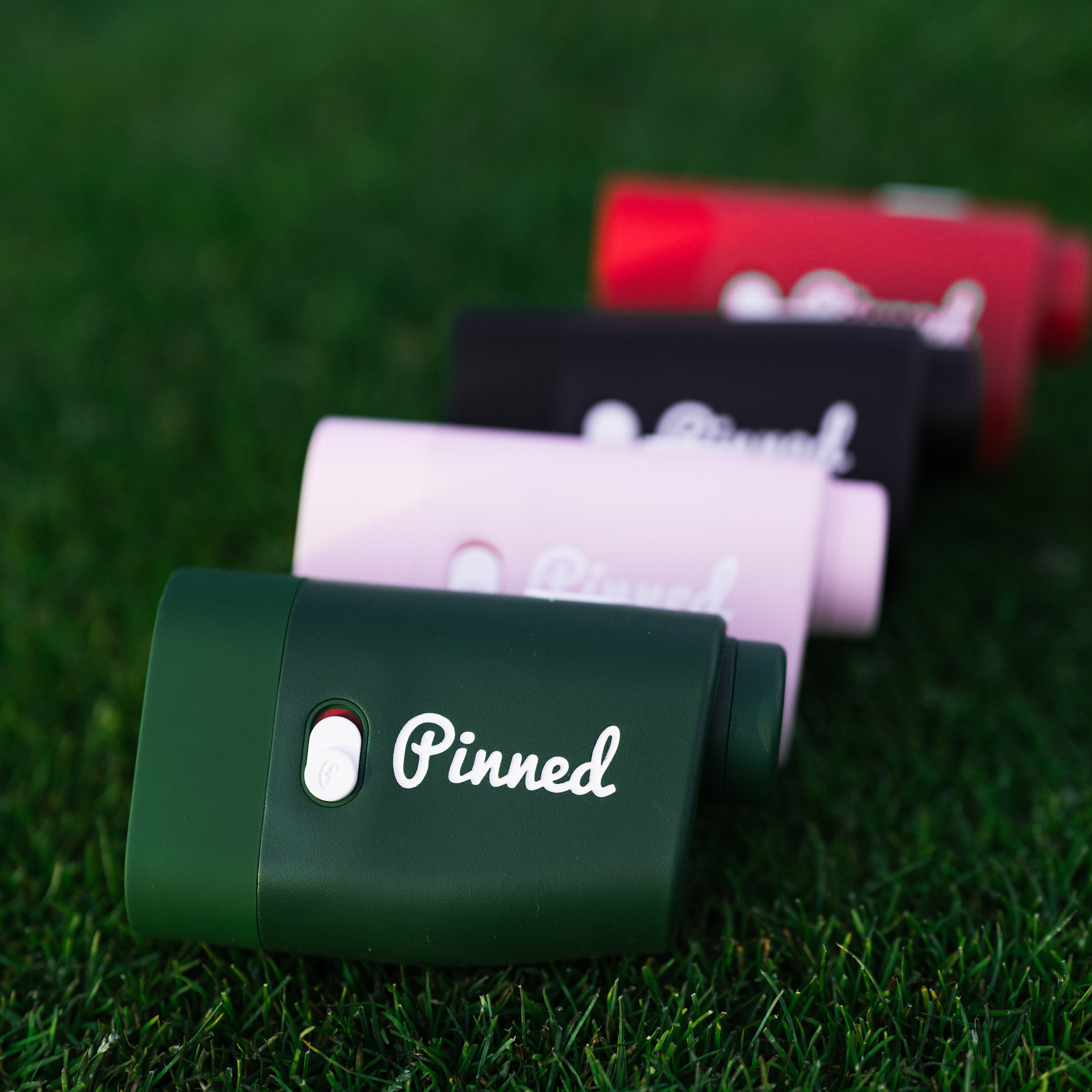 Four rangefinders of various colors with the word 'Pinned' in white as branding and placed on a golf green