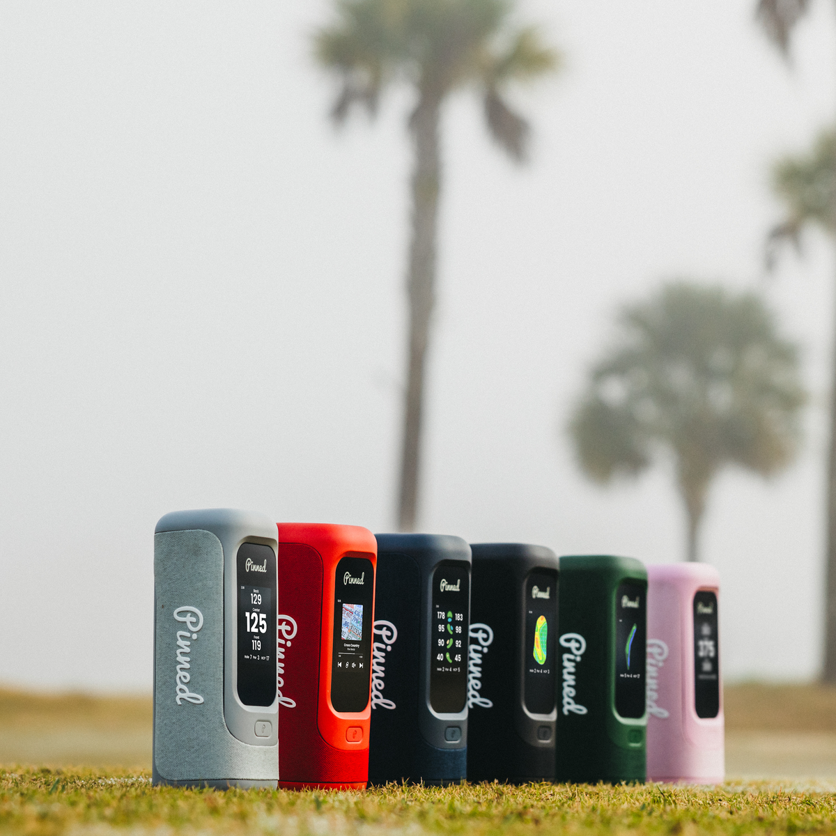 Portable speakers of various colors with the 'Pinned' branding in white lined up on a golf course