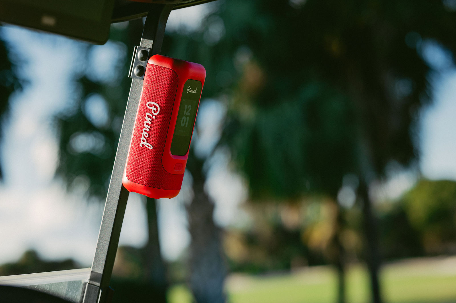 A red portable speaker with a digital display screen, magnetically attached on the black metal frame of a golf cart outdoors on a blurred golf course background.
