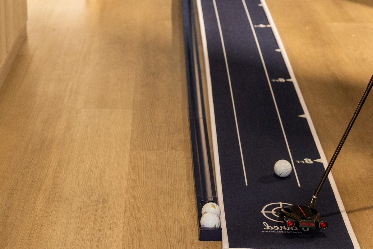 A closeup of a blue 'Pinned Golf' putting mat with a golf club and golf ball setup on a wooden floor.