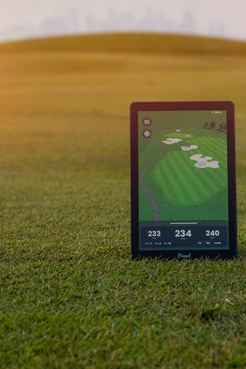 A 'Pinned Golf' branded caddie GPS showcasing a screen and standing upright on a grassy field, with a blurred golf course landscape in the background at sunset.