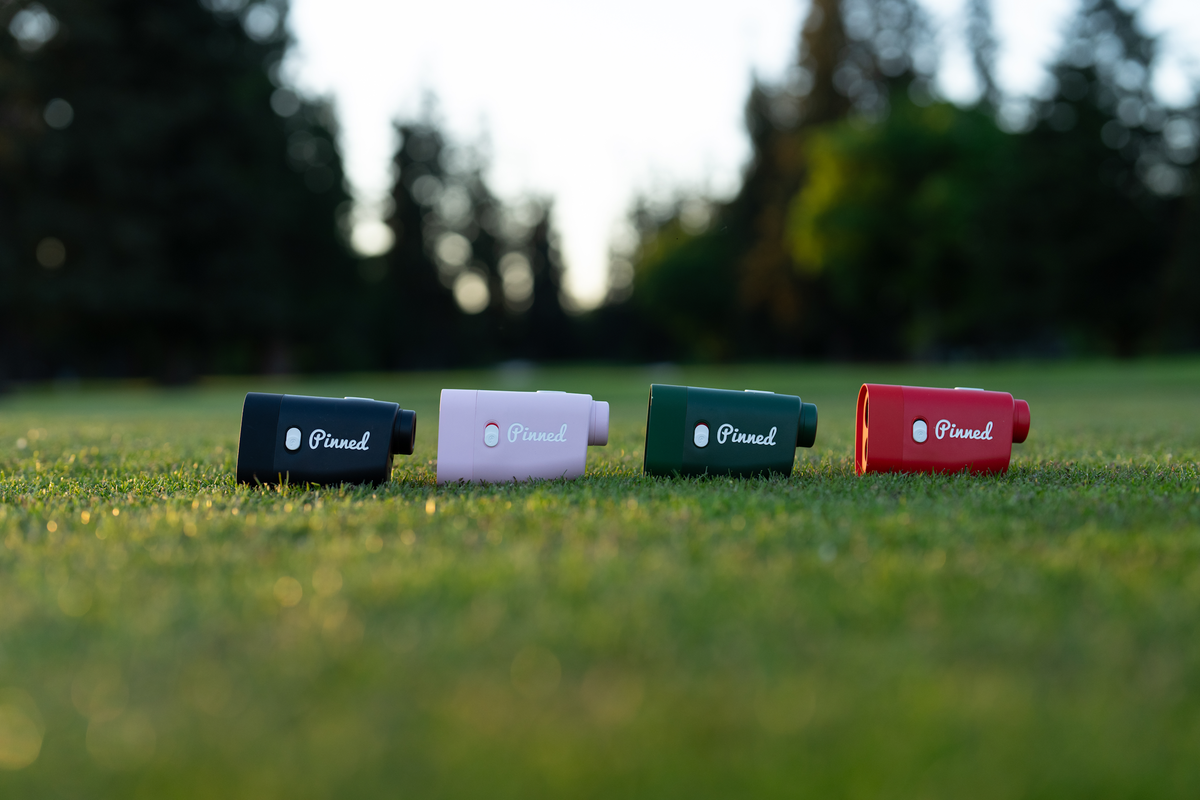 Set of four 'Pinned Golf' Prism+ Rangefinders on a golf course setting with blurred trees in the background.