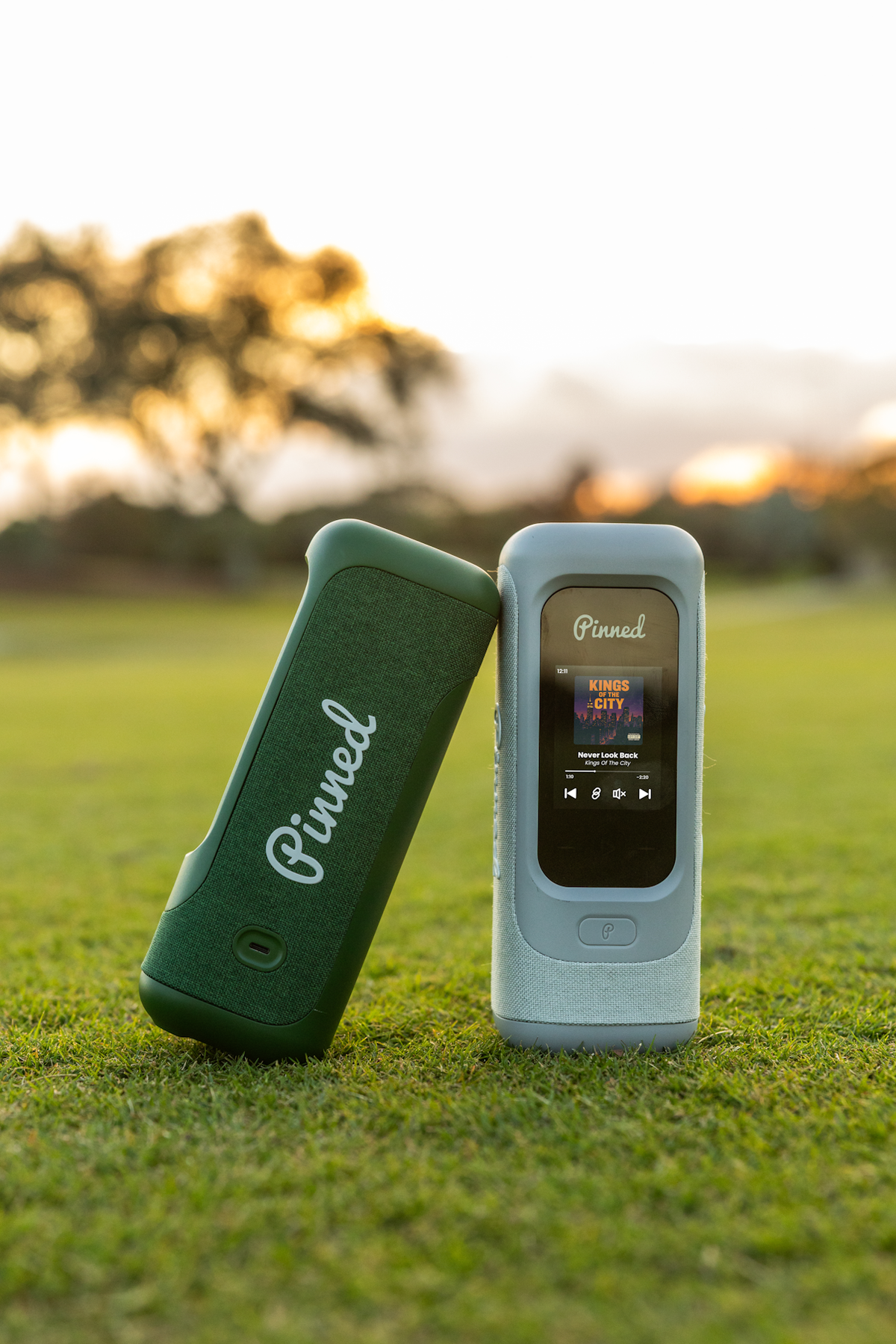 A set of two 'Pinned Golf' branded portable speakers on a golf course setting with blurred trees in the background.
