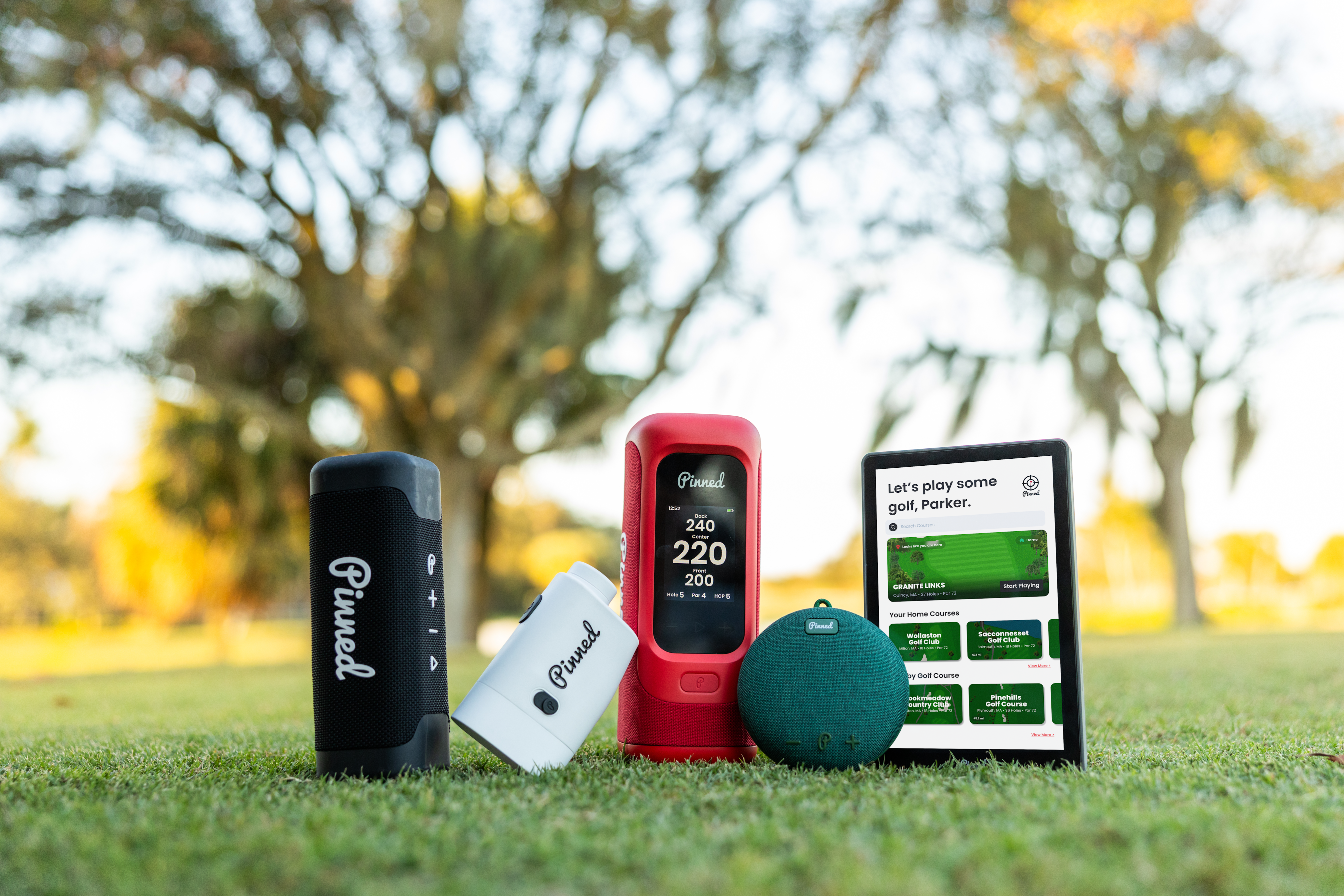 A set of 'Pinned Golf' brand technology accessories arranged on grass on a golf course setting with blurred trees in the background. The set includes a black sound stick speaker, a white prism+ rangefinder, a red sound stick pro portable speaker showing a measurement on screen, a green dart 2.0 speaker, and a caddie GPS smart tablet displaying a golf course app.