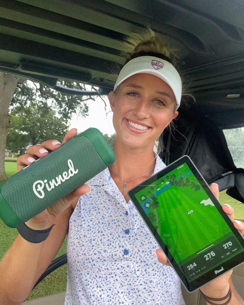 A woman, Hunter Nugent, in a golf cart holds a green Pinned portable speaker in one hand and a GPS smart tablet in the other, with trees and grass visible in the background.