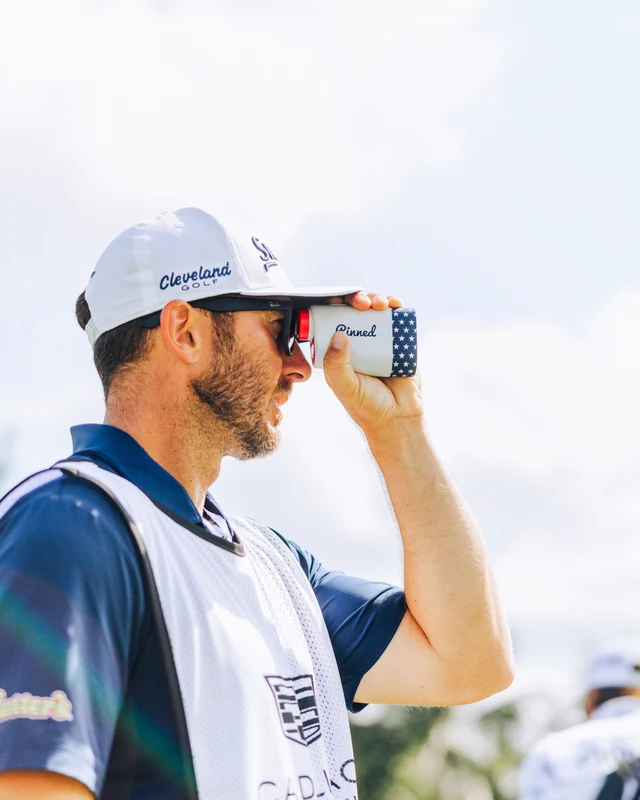A man, Jeff Hamley, wearing a Cleveland golf cap and sunglasses, holding a rangefinder to his eye with a blurred background.