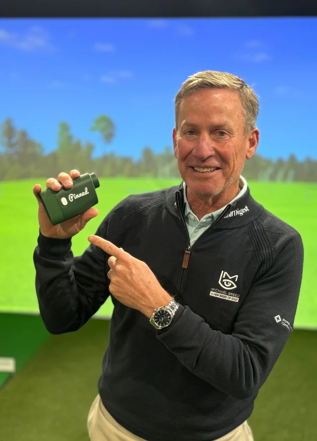 A man, Michael Breed, in a golf simulator setting holding a device and pointing at it.