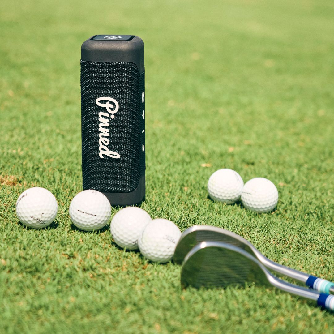 Navy blue portable speaker with 'Pinned' branding in white on a golf course with golf clubs and balls