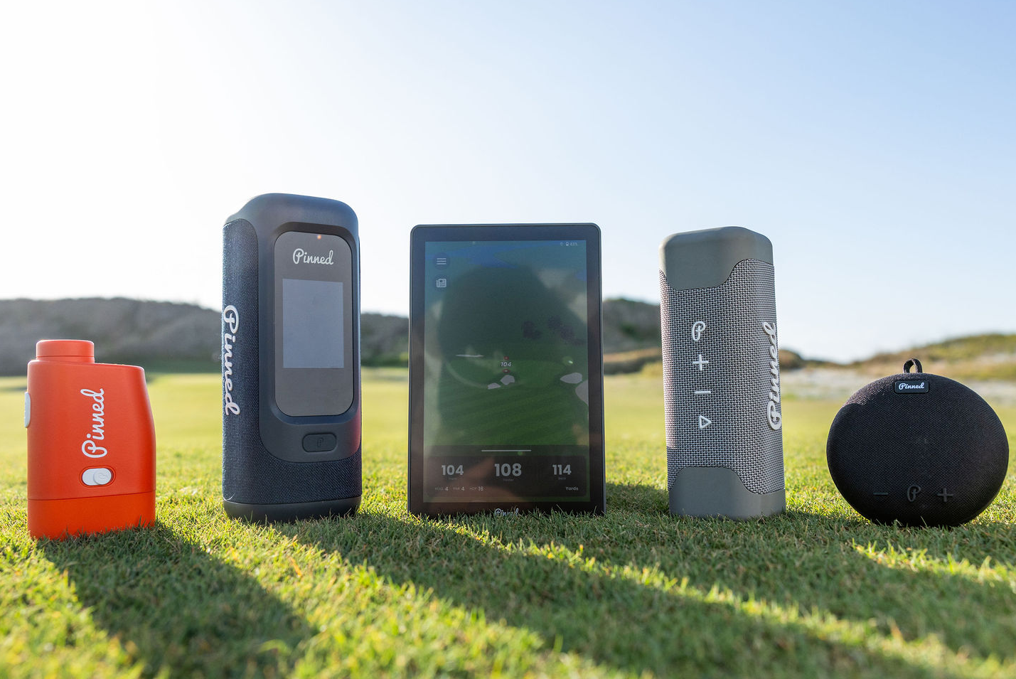 A lineup of five 'Pinned Golf' products setup on a golf course. From left to right: an orange rangefinder, a blue rectangular portable speaker with an integrated screen, a caddie GPS smart tablet, a grey rectangular portable speaker, a black round portable speaker.