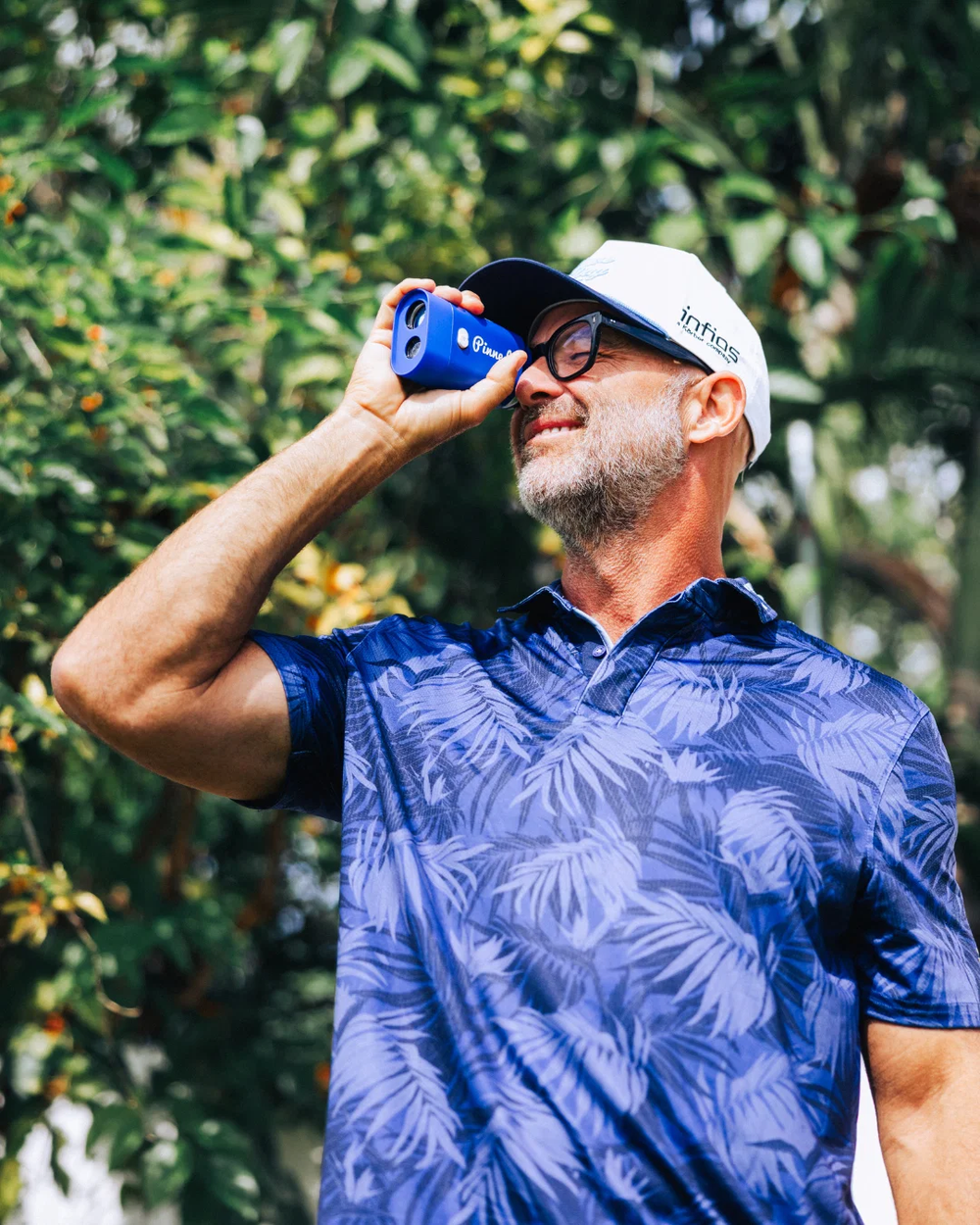 A man, Scott Vail, wearing a blue and white palm leaf patterned polo shirt and a white baseball cap, holding a blue rangefinder outdoors in front of green foliage.